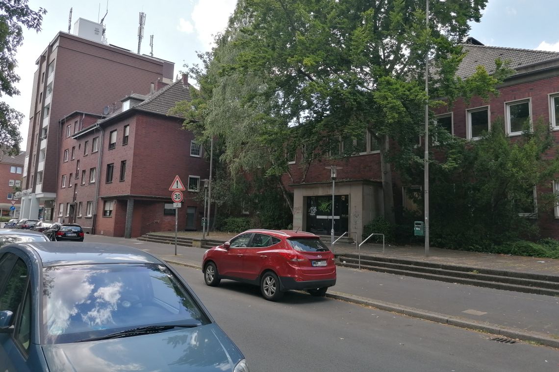 Herne: Bürger kämpfen für das Hallenbad Eickel – Politik ignoriert Warnzeichen Ehemaliges Hallenbad Eickel aus rotem Klinker in innerstädtischer Lage, mit graffitibeschmierter Eingangstür, umgeben von parkenden Autos, Bäumen und Gehweg – Straßenszene mit Tempo-30-Schild im Vordergrund.