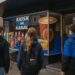 Drei Jugendliche mit Rucksäcken stehen vor dem Kiosk am Kanal mit Werbeplakaten für Snacks und Getränke.