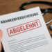 Abgelehnt von der Krankenkasse? So wehren Sie sich erfolgreich! Nahaufnahme eines Ablehnungsschreibens einer Krankenversicherung mit dem deutlich sichtbaren roten Stempel ‚ABGELEHNT‘. Im Hintergrund liegen ein Stethoskop und ein schwarzer Kugelschreiber auf einem Holztisch, eine Hand hält das Schreiben fest.