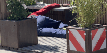 Eine alkoholisierte Person liegt ausgestreckt auf dem Boden einer hölzernen Stadtterrasse in Herne, umgeben von Pflanzkübeln; die Szene wirkt verlassen und still, das Umfeld zeigt keine Passant:innen.