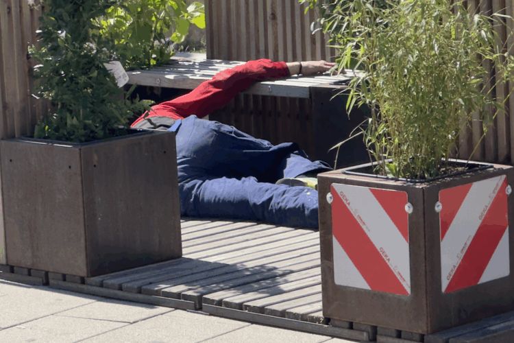 Eine alkoholisierte Person liegt ausgestreckt auf dem Boden einer hölzernen Stadtterrasse in Herne, umgeben von Pflanzkübeln; die Szene wirkt verlassen und still, das Umfeld zeigt keine Passant:innen.