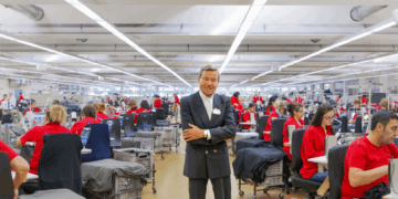 Wolfgang Grupp steht inmitten einer Trigema-Näherei, umgeben von Mitarbeiterinnen an Nähmaschinen in roter Arbeitskleidung.