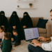 Muslimisches Wohnzimmer mit drei Frauen im Niqab auf dem Sofa, spielenden Kindern auf dem Teppich und einem Mann, der am Laptop die Webseite eines islamischen Heiratsportals aufruft.