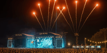 Menschenmenge beim San Hejmo Festival bei Nacht, beleuchtete Bühne mit Feuerwerk und Getränkeständen im Hintergrund.