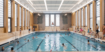 Modernisiertes Hallenbad Eickel mit vielen Schwimmer:innen im Becken, Holzverkleidung an den Wänden und original erhaltenen Wandmosaiken.