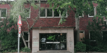 Ehemaliges Hallenbad Eickel in Herne aus rotem Klinker mit starkem Sanierungsbedarf in einer deutschen Innenstadt. Der Eingangsbereich ist von Graffiti verunstaltet, Bäume wachsen direkt an der Fassade. Das Gebäude wirkt verlassen und ungepflegt. Straßenschild mit Tempo-30-Zone im Vordergrund.