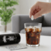 Eine Hand hält eine weiße Tablette über einem Glas Cola mit Eiswürfeln. Daneben liegt eine leere Afri-Cola-Dose und ein Blister mit Tabletten.