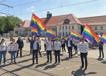 Der Schein trügt: Soll der Heinz-Rühmann-Platz in Herne nach NS-Verstrickungen weichen? Eine Gruppe von Personen steht auf dem Heinz-Rühmann-Platz vor dem Hauptbahnhof Wanne-Eickel und hält Regenbogenfahnen sowie Schilder mit den Aufschriften "TOLERANZ & LIEBE FÜR HERNE" und "JÜRGEN MARCUS PLATZ". Die Szene findet unter blauem Himmel statt und zeigt eine Versammlung zur Unterstützung der LGBTQ+-Community und zur Würdigung des Jürgen Marcus Platzes.