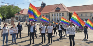 Eine Gruppe von Personen steht auf dem Heinz-Rühmann-Platz vor dem Hauptbahnhof Wanne-Eickel und hält Regenbogenfahnen sowie Schilder mit den Aufschriften "TOLERANZ & LIEBE FÜR HERNE" und "JÜRGEN MARCUS PLATZ". Die Szene findet unter blauem Himmel statt und zeigt eine Versammlung zur Unterstützung der LGBTQ+-Community und zur Würdigung des Jürgen Marcus Platzes.