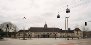 Hauptbahnhof Wanne-Eickel (Herne) mit zwei Kabinen der urbanen Seilbahn, die über das Bahnhofsgebäude und den leeren Vorplatz schweben. Visualisierung des umstrittenen ÖPNV-Projekts General Blumenthal XI.