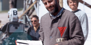 Jerry Lewis († 91) während der Dreharbeiten zu „The Day the Clown Cried“: graue Häftlingskleidung mit rotem, auf der Spitze stehendem Stoffdreieck als Kennzeichnung politischer Häftlinge im nationalsozialistischen Konzentrionslager-System; Klemmbrett in der Hand, Filmset im Hintergrund.