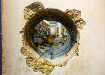 Dokumentation der Einbruchsmethode: Ein präzise gebohrtes Loch in einer dicken Stahlbetonwand gewährt Einblick in den dahinterliegenden Tresorbereich mit Regalen. Sichtbare Bohrspuren an den Innenwänden des Durchbruchs sowie gelbliche Isolations- oder Rückstände an der Außenseite.