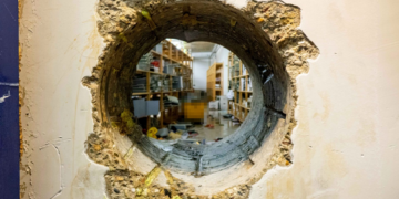 Dokumentation der Einbruchsmethode: Ein präzise gebohrtes Loch in einer dicken Stahlbetonwand gewährt Einblick in den dahinterliegenden Tresorbereich mit Regalen. Sichtbare Bohrspuren an den Innenwänden des Durchbruchs sowie gelbliche Isolations- oder Rückstände an der Außenseite.