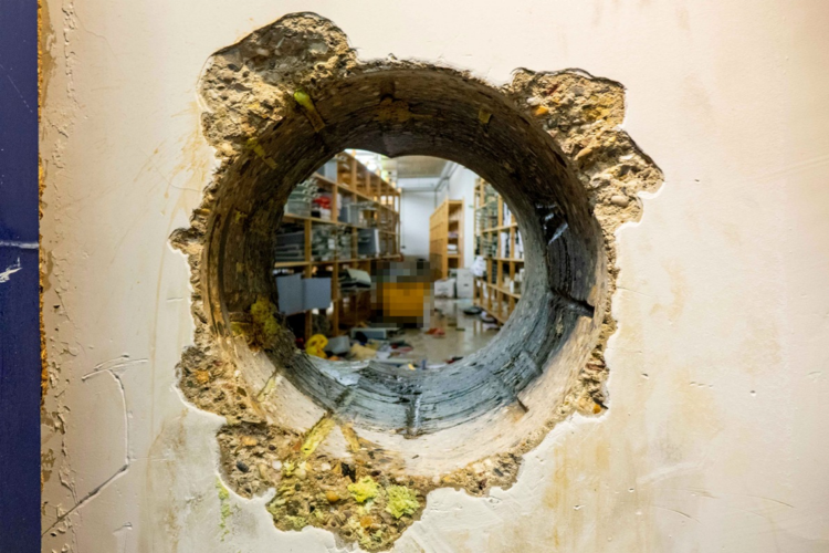 Dokumentation der Einbruchsmethode: Ein präzise gebohrtes Loch in einer dicken Stahlbetonwand gewährt Einblick in den dahinterliegenden Tresorbereich mit Regalen. Sichtbare Bohrspuren an den Innenwänden des Durchbruchs sowie gelbliche Isolations- oder Rückstände an der Außenseite.
