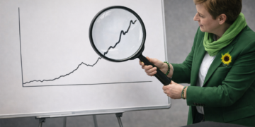 Politiker:in in Grün hält eine große Lupe über einen kleinen Abschnitt einer unbeschrifteten Kurve auf einer Tafel, neutraler Besprechungsraum.