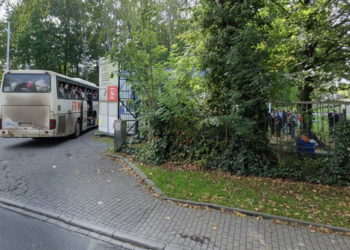 Ein vollbesetzter Reisebus mit Geflüchteten fährt in die Einfahrt einer Aufnahmeeinrichtung; Menschen warten hinter einem Zaun.
