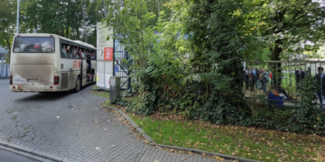 Ein vollbesetzter Reisebus mit Geflüchteten fährt in die Einfahrt einer Aufnahmeeinrichtung; Menschen warten hinter einem Zaun.