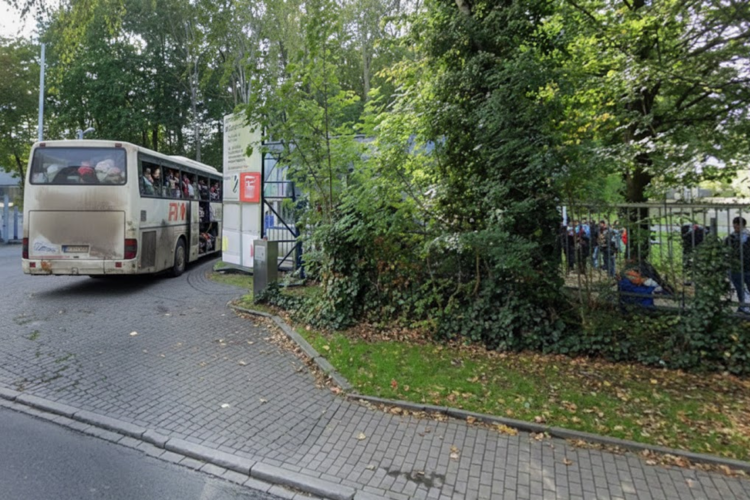 Ein vollbesetzter Reisebus mit Geflüchteten fährt in die Einfahrt einer Aufnahmeeinrichtung; Menschen warten hinter einem Zaun.