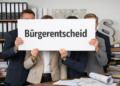 Anonyme Gruppe in Businesskleidung hält ein Schild mit „Bürgerentscheid“ vor Aktenordnern, Bauplänen und Stadtplan in einem Büro.