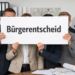 Anonyme Gruppe in Businesskleidung hält ein Schild mit „Bürgerentscheid“ vor Aktenordnern, Bauplänen und Stadtplan in einem Büro.