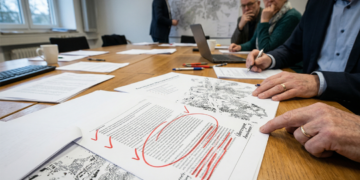 Nahaufnahme eines Entwurfs zur kommunalen Wärmeplanung in Herne mit roten Korrekturen und kritischen Anmerkungen. Im Hintergrund sitzen Personen an einem Konferenztisch bei einer Fachdiskussion über Wärmenetze und Energieversorgung.