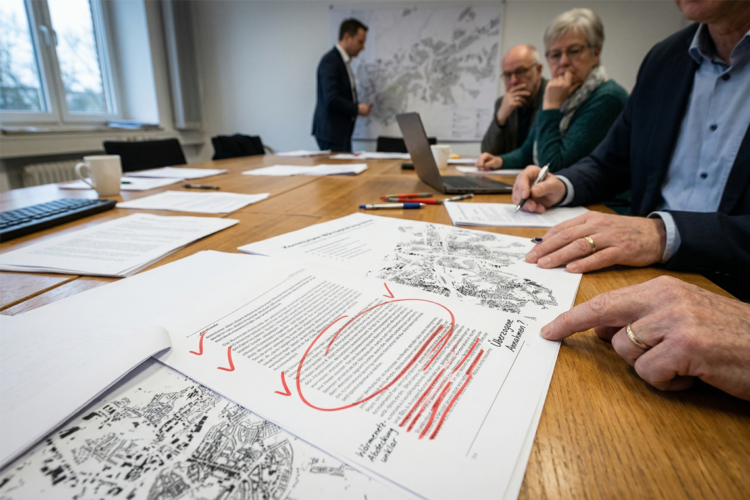Nahaufnahme eines Entwurfs zur kommunalen Wärmeplanung in Herne mit roten Korrekturen und kritischen Anmerkungen. Im Hintergrund sitzen Personen an einem Konferenztisch bei einer Fachdiskussion über Wärmenetze und Energieversorgung.
