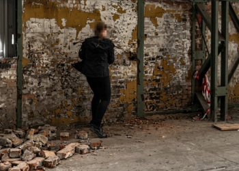 Eine Frau in einem schwarzen Kapuzenpullover steht mit dem Rücken zur Kamera auf dem Funkenbergareal in Herne, einer Abrisshalle für das n8werk Event, und hält ein Werkzeug vor eine marode, gelb-weiße Ziegelwand.