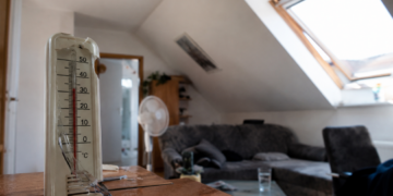Innenansicht einer aufgeheizten Dachgeschosswohnung mit Dachschrägen, Thermometer ohne lesbare Werte und starkem Sonnenlichteinfall.