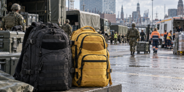Ein schwarzer und ein gelber Notfallrucksack stehen im Vordergrund einer Hamburger Hafen- und Logistikszene; im Hintergrund befinden sich Militärfahrzeuge, anonyme Soldat:innen und Einsatzkräfte vor der Stadtkulisse.