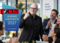 stefanbuddesiegel als Leading Innovator 2027 ausgezeichnet Mann im schwarzen Anzug und mit Brille steht in einem modernen Büro vor applaudierenden Kolleg:innen und hebt die Faust. Links ist das Logo „FOCUS Leading Innovator 2027“ eingeblendet.