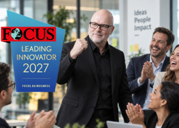 stefanbuddesiegel als Leading Innovator 2027 ausgezeichnet Mann im schwarzen Anzug und mit Brille steht in einem modernen Büro vor applaudierenden Kolleg:innen und hebt die Faust. Links ist das Logo „FOCUS Leading Innovator 2027“ eingeblendet.