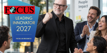 Mann im schwarzen Anzug und mit Brille steht in einem modernen Büro vor applaudierenden Kolleg:innen und hebt die Faust. Links ist das Logo „FOCUS Leading Innovator 2027“ eingeblendet.