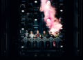 Rammstein „Live in Mexico City“: Konzertfilm ab November Dramatisches Pressebild der Band Rammstein (ca. 2026), das die sechs Mitglieder in Kostümen auf einer dunklen, vertikalen Industrie-Bühnenkonstruktion zeigt. Sie stehen hinter einer beleuchteten Brüstung mit leuchtenden Kreuzsymbolen. Über ihnen steigt eine dichte Wolke aus rosa-weißem Pyro-Rauch auf, beleuchtet von mehreren kühlen Bühnenscheinwerfern, die eine düstere, mechanische Ästhetik erzeugen.