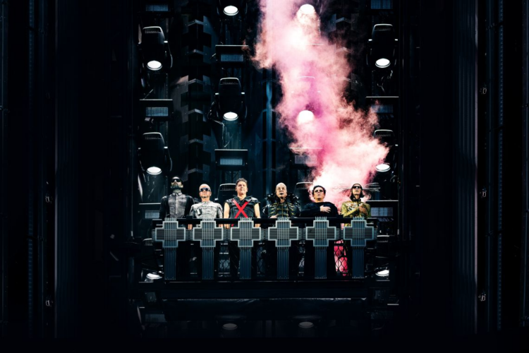 Rammstein „Live in Mexico City“: Konzertfilm ab November Dramatisches Pressebild der Band Rammstein (ca. 2026), das die sechs Mitglieder in Kostümen auf einer dunklen, vertikalen Industrie-Bühnenkonstruktion zeigt. Sie stehen hinter einer beleuchteten Brüstung mit leuchtenden Kreuzsymbolen. Über ihnen steigt eine dichte Wolke aus rosa-weißem Pyro-Rauch auf, beleuchtet von mehreren kühlen Bühnenscheinwerfern, die eine düstere, mechanische Ästhetik erzeugen.