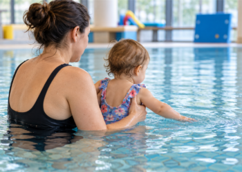 Eine erwachsene Person hält ein Kleinkind im Hallenbad, während es mit der Hand das Wasser berührt.