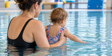 Eine erwachsene Person hält ein Kleinkind im Hallenbad, während es mit der Hand das Wasser berührt.