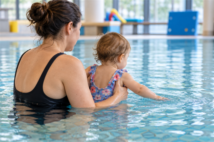 Eine erwachsene Person hält ein Kleinkind im Hallenbad, während es mit der Hand das Wasser berührt.