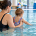 Eine erwachsene Person hält ein Kleinkind im Hallenbad, während es mit der Hand das Wasser berührt.