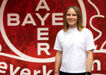 Michelle Böhmke übernimmt im Sommer 2027 die Leitung des Hauses der Talente von Bayer 04 Leverkusen und steht vor einer roten Vereinswand.